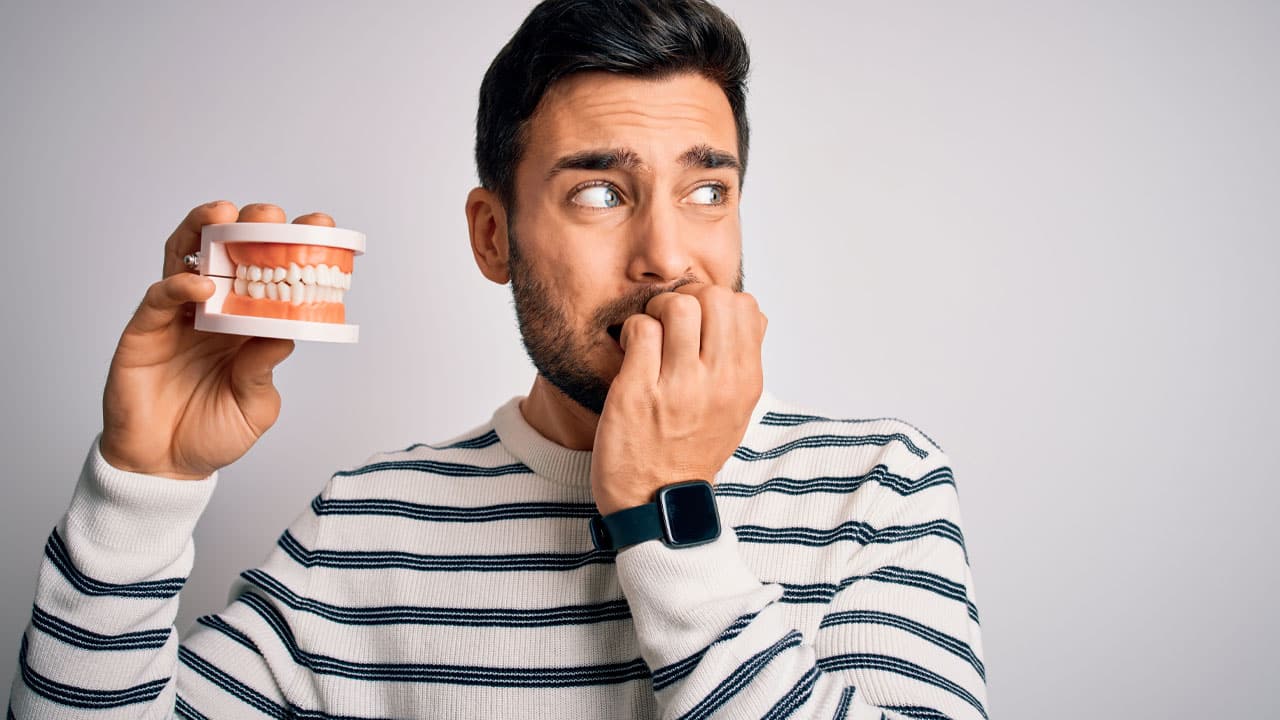 Young handsome man with beard holding plastic denture teeth over white background looking stressed and nervous with hands on mouth biting nails
